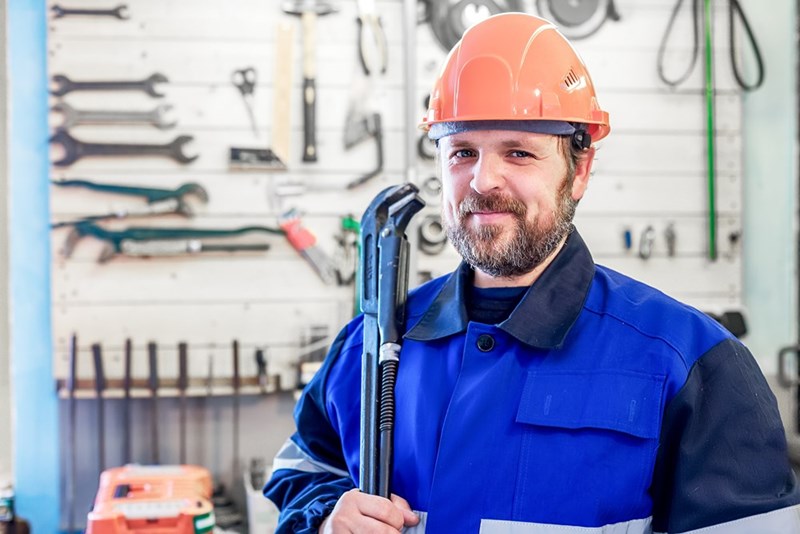 Smiling worker in an orange hard hat and blue uniform holding a large wrench in a tool-lined workshop.