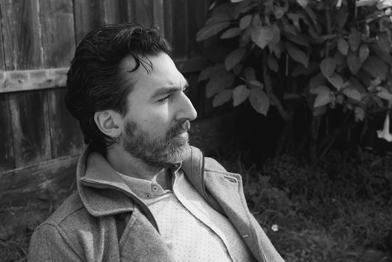 Black-and-white side-profile portrait of a bearded man sitting outdoors beside a wooden fence and leafy plants.