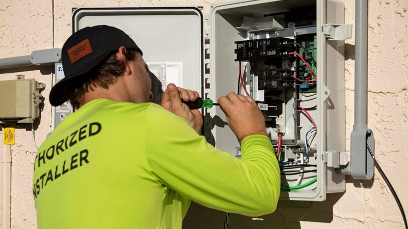 An electrician with a backwards hat and a hi-vis shirt works on an electrical box outside of someone's home (stock).