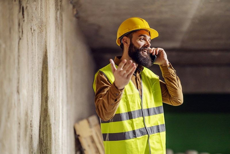 A blue-collar tradesman wearing a hard hat and hi-vis vest talks on a cellphone while smiling (model).