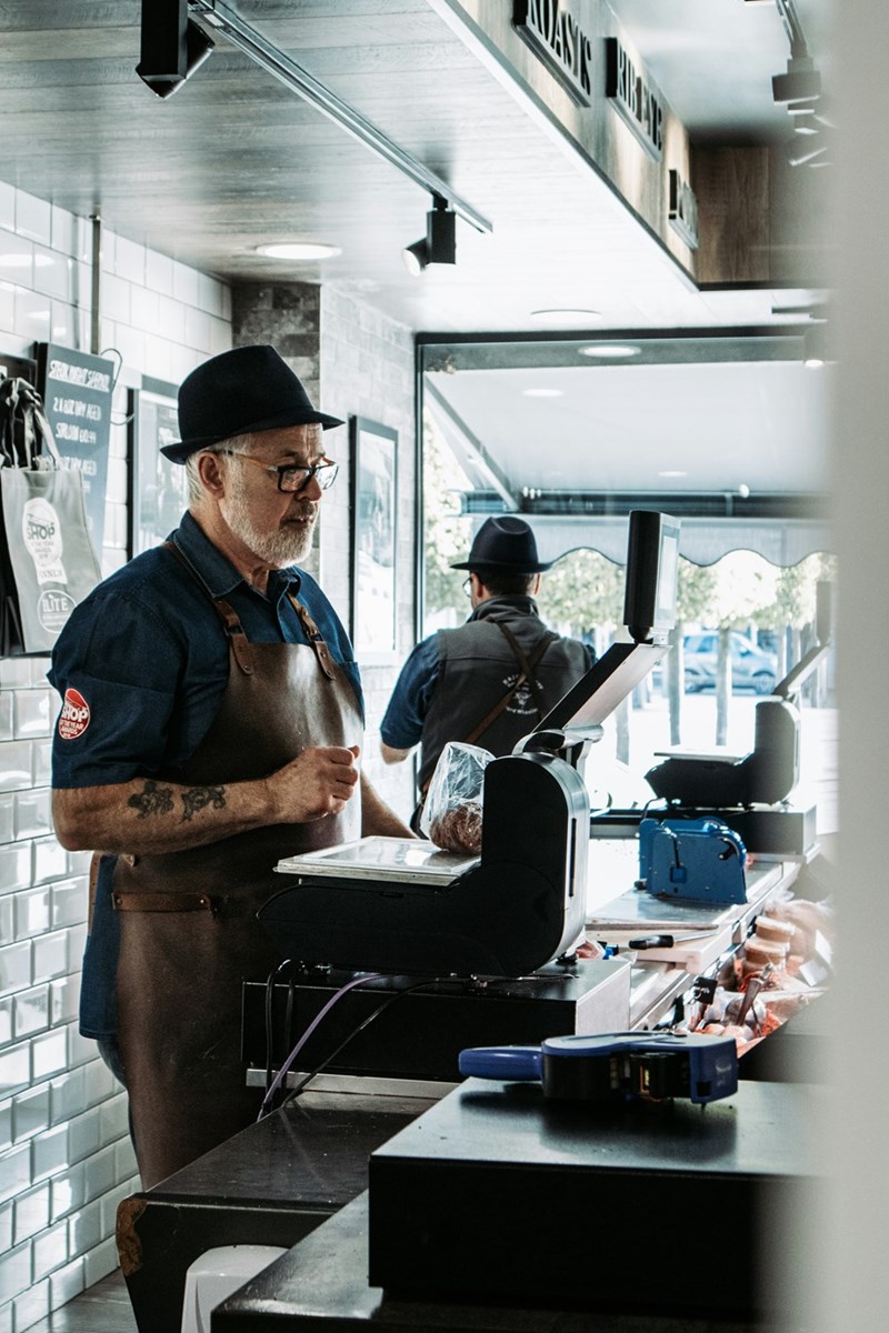 Man wearing a hat and leather apron standing at a deli counter beside a register and scale.