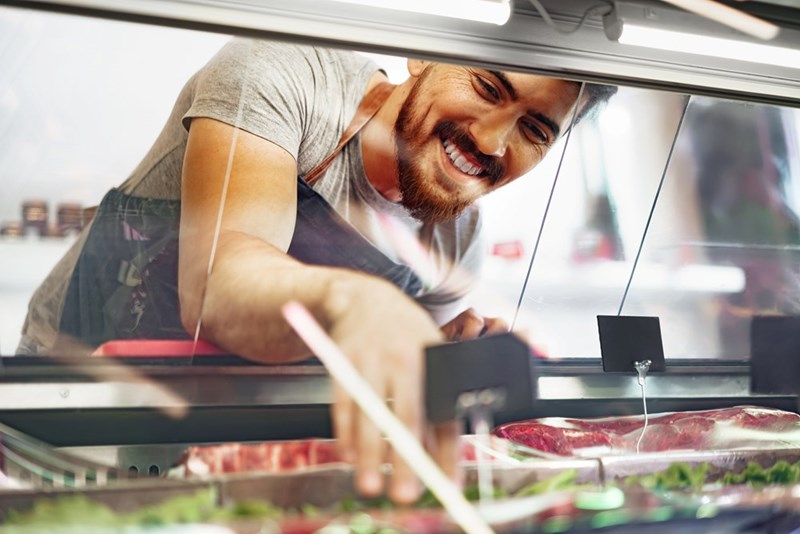 Smiling man leaning over a refrigerated meat display and reaching behind the counter.
