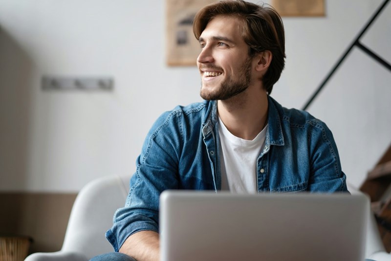 Smiling man in a denim shirt sits indoors behind a laptop, looking off to the side.