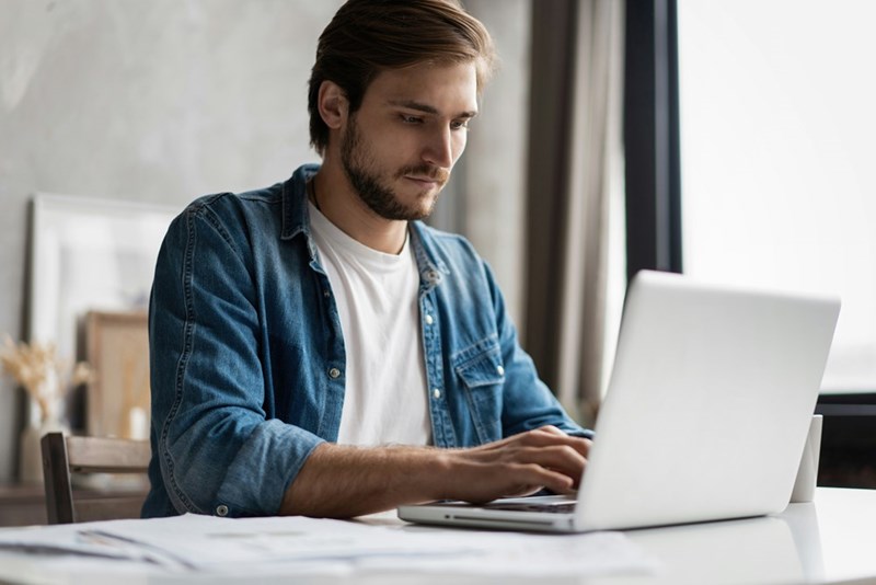 Man in a denim shirt typing on a laptop at a table near a window.