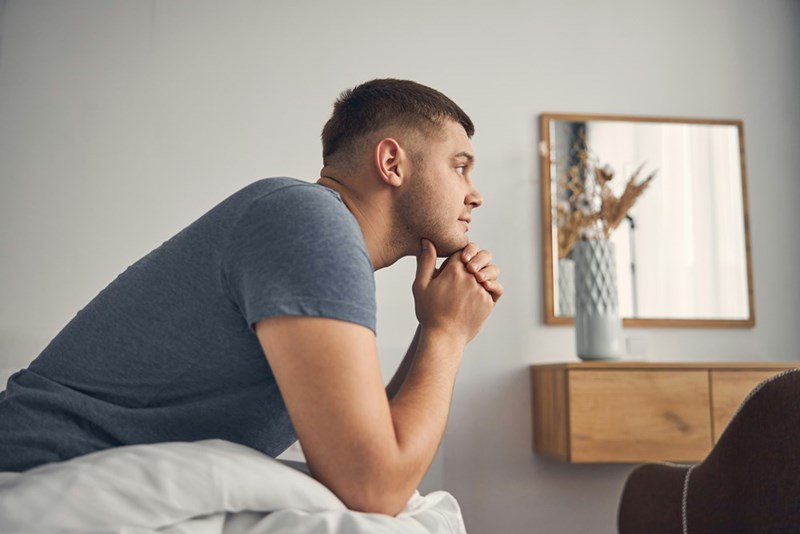 Man in a gray shirt leans on a bed with hands under his chin in a bright bedroom.