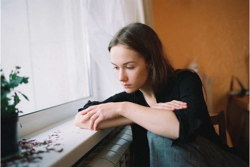 Woman leaning on a windowsill indoors with folded arms and a sad, thoughtful expression.