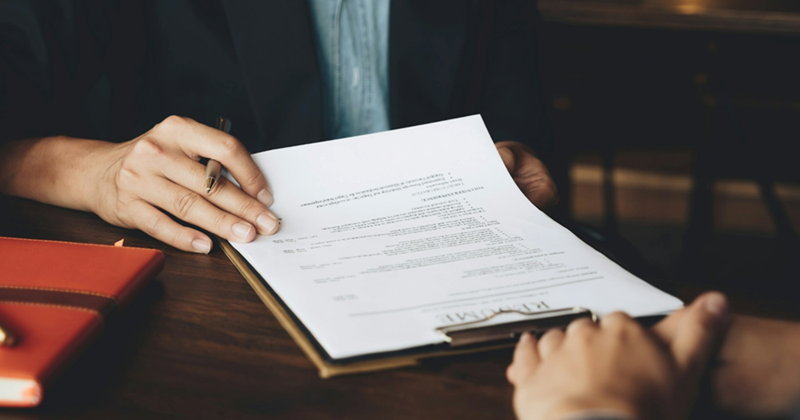 A woman's hand flicking through a resume on a clipboard as somebody sits opposite them