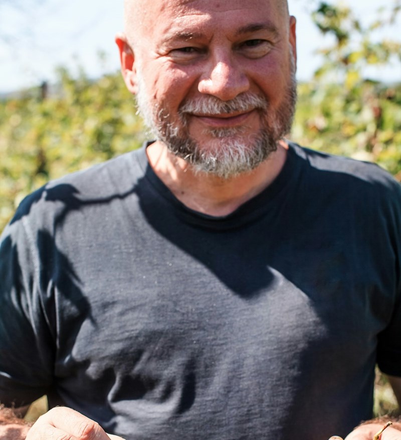 Man with a gray beard smiles outdoors in a black shirt with greenery in the background.