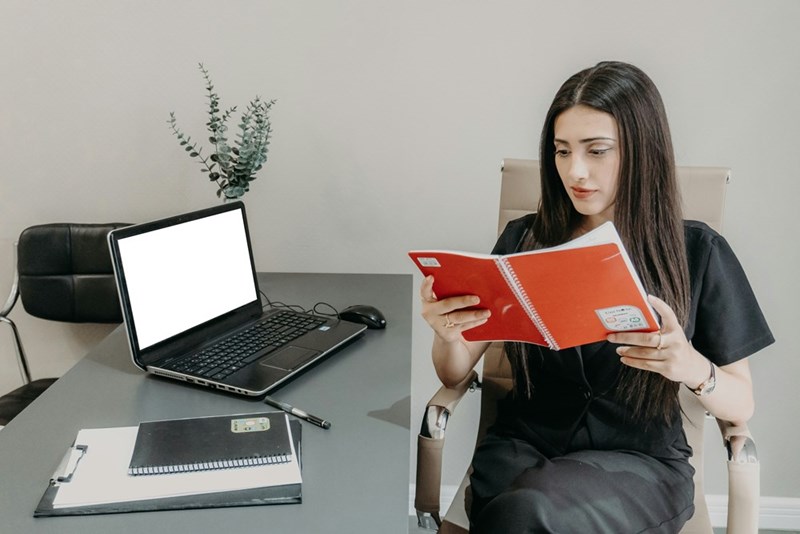 woman reading a book at a desk with a laptop