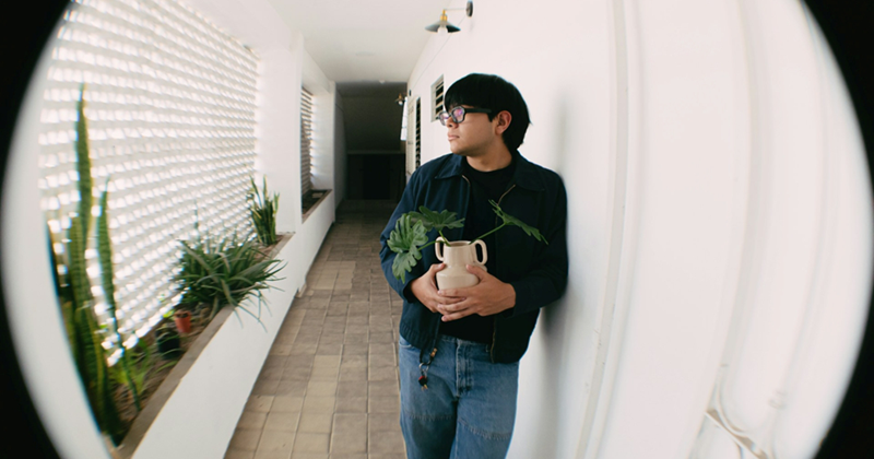 A man leaning in a hallway outside an apartment holding a plant, seen through a fisheye lens