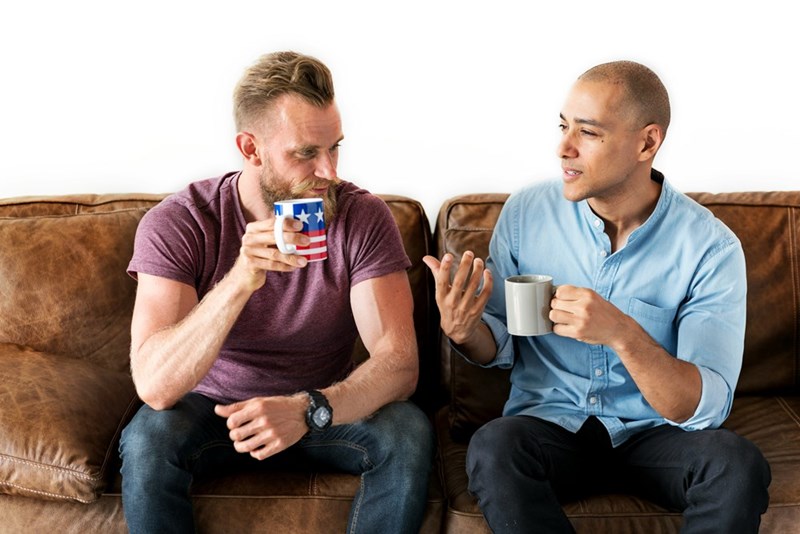 Two men sitting on a leather brown couch holding coffee mugs and talking.