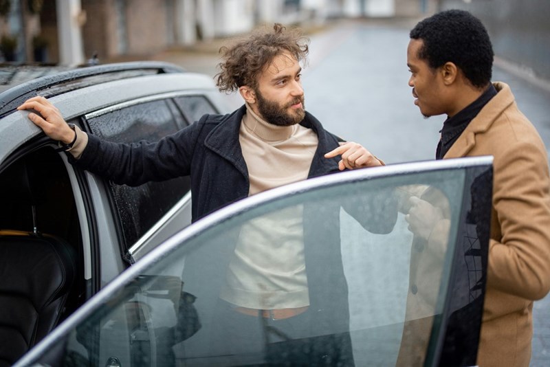 Two men talking on the streets right beside a car.