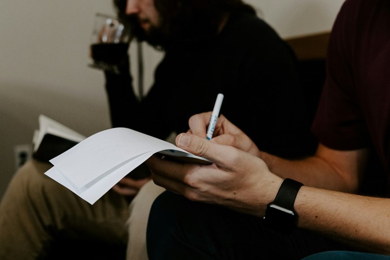 Man writing in a notebook besides other man sitting next to him.