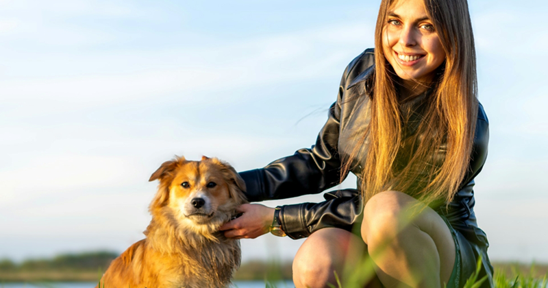 A young woman smiles as she kneels and pets a small, long-haired dog beside her outside