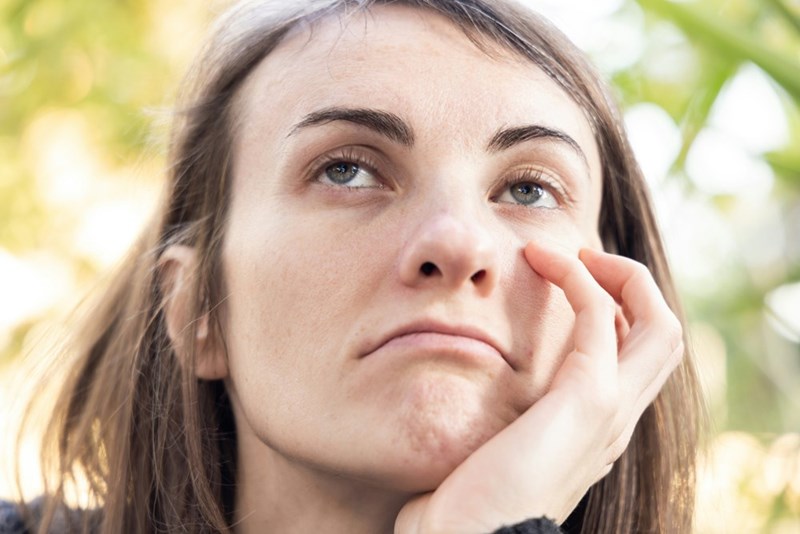 Woman resting her cheek on her hand looks upward with a bothered expression against a blurred green background.