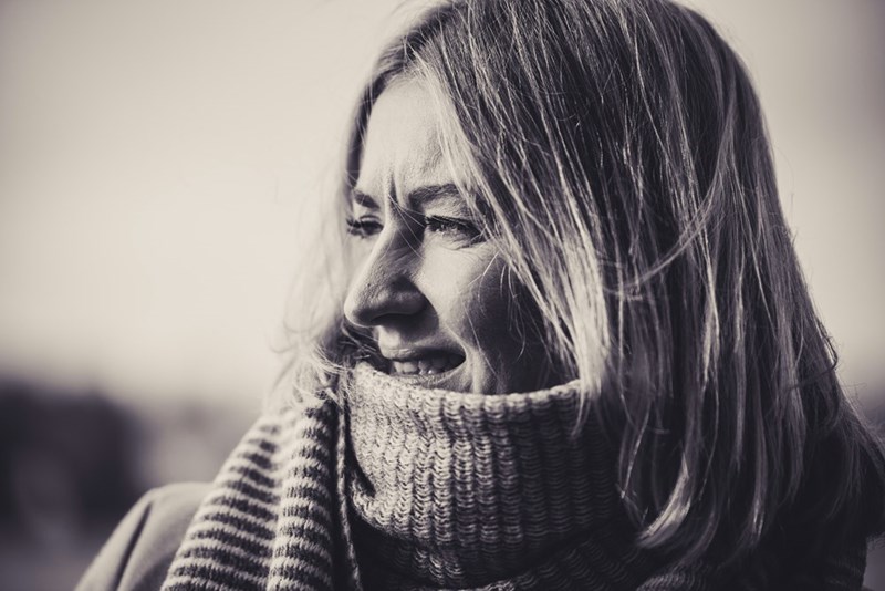 Woman in a thick scarf looks to the side with a conflicted, cornered expression in a black-and-white close-up.