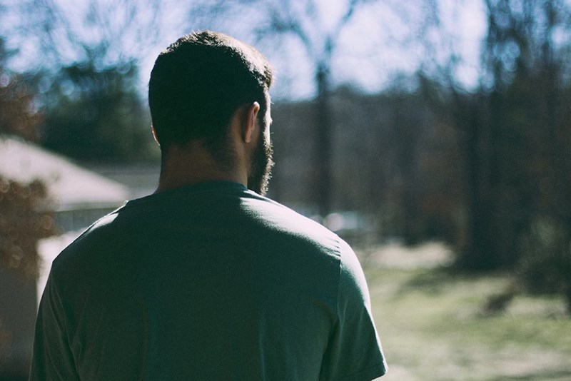 Man in a green shirt seen from behind standing outdoors in bright sunlight with blurred trees ahead.