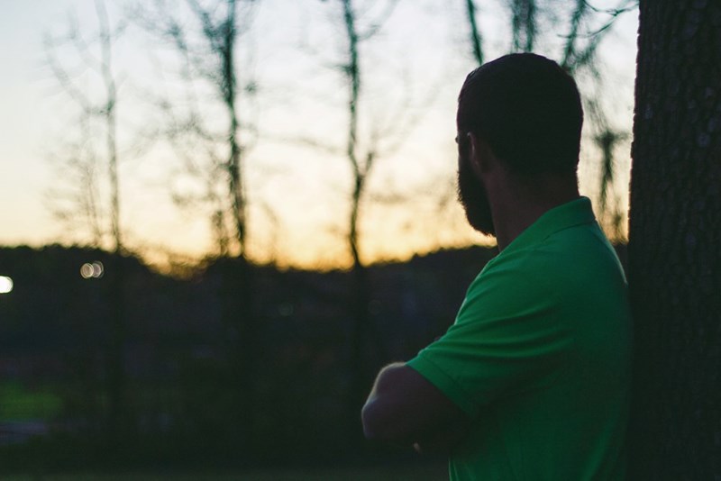Man in a green shirt stands by a tree at sunset, seen from behind with bare trees in the background.