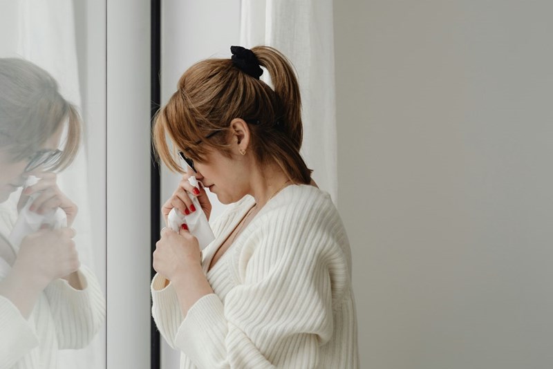 Woman standing by a window while holding a tissue.