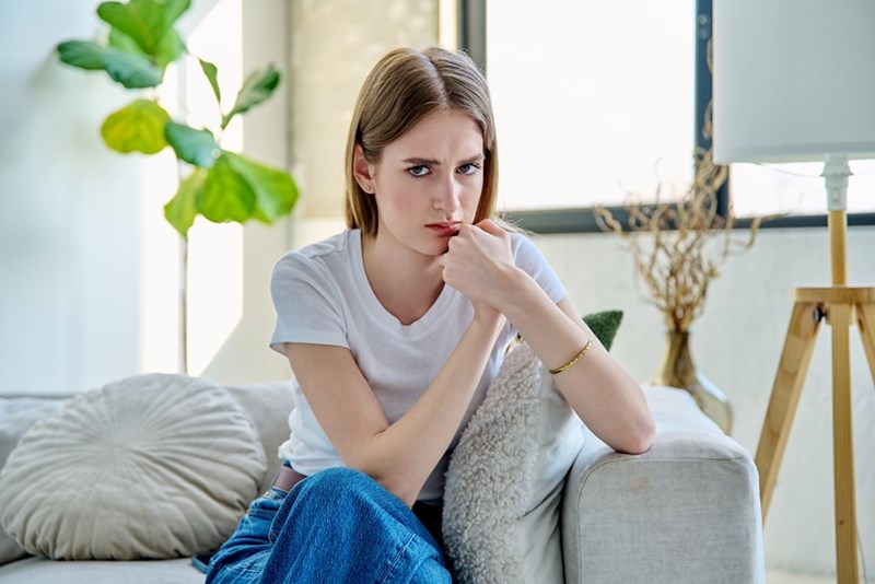 Young woman sitting on a couch with her hand near her mouth, looking tense in a bright living room.