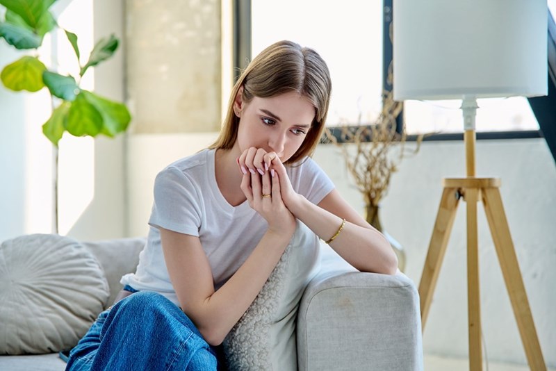 Young woman sitting on a couch with clasped hands, looking worried in a bright living room.