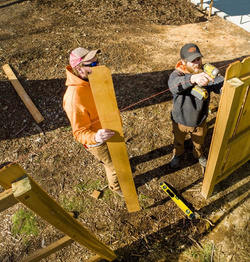 Two workers installing a wooden fence with a drill, string line, level, and disturbed soil in a sunny yard.