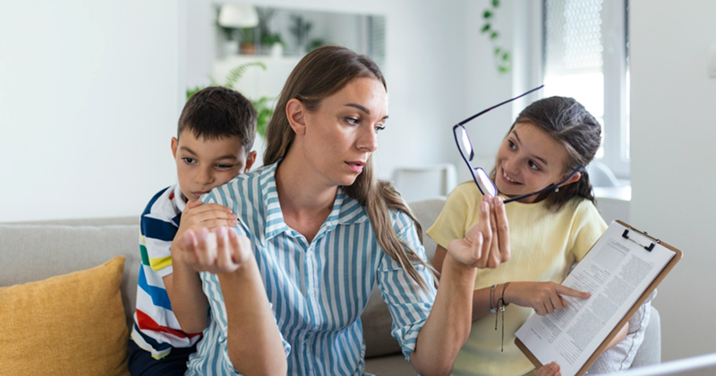 A representation of a mom holding glasses and shrugging while sitting on a couch, with two kids on either side of her