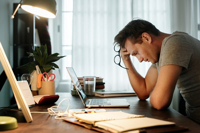 Man leaning over a laptop at a desk, holding glasses and looking stressed under a desk lamp.