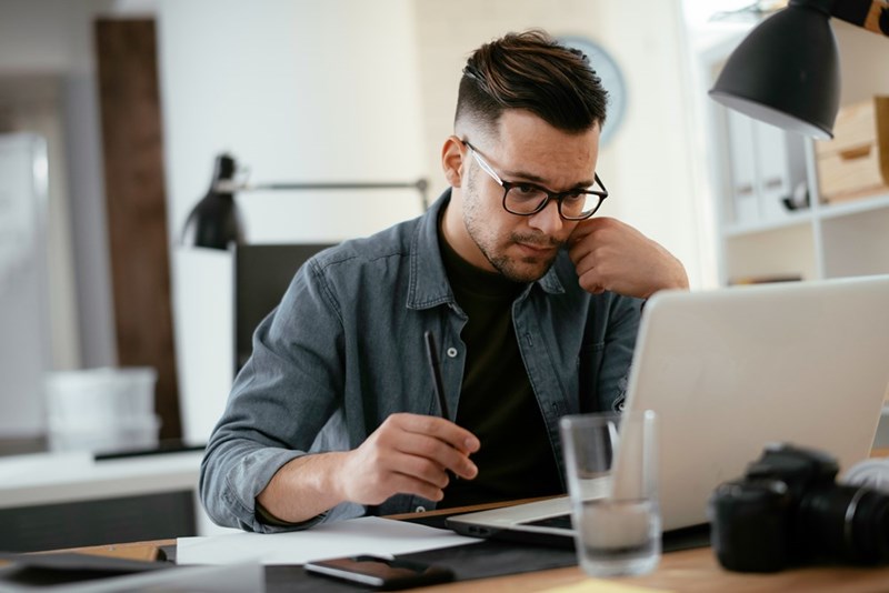 Man wearing glasses works at a desk with a laptop, pen, glass of water, and desk lamp nearby.