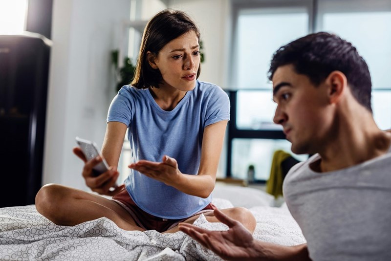 Woman holding a phone with a hurt, questioning expression, while talking to a man on a bed in a bright room.