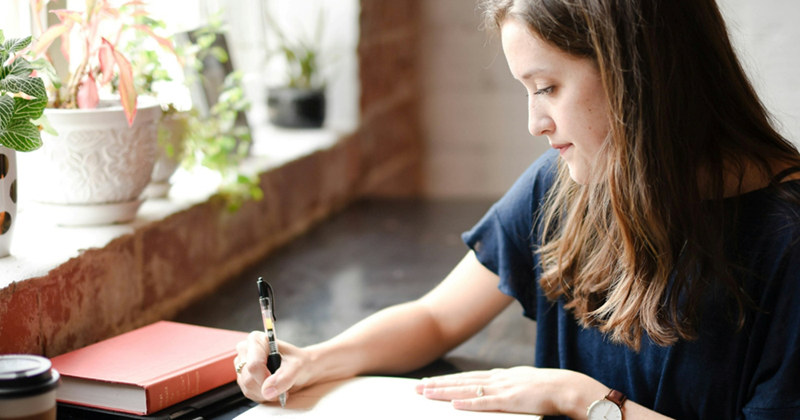 A representation of a woman sitting in a coffee shop, writing something in a notebook with other books beside her