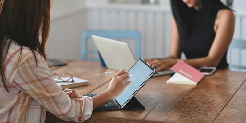 Two women working while sitting in front of each other with a computer and a laptop, depicted by models.