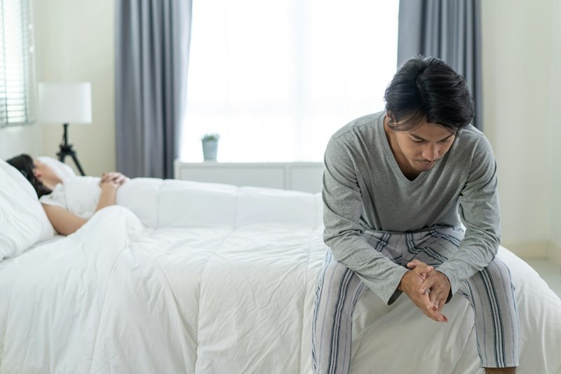 Man sitting on the edge of a bed looking worried while his partner rests in the background.