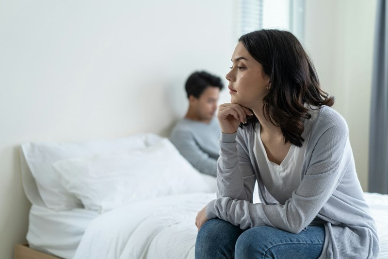Pensive woman looking away while sitting near a bed, with her partner out of focus behind her.