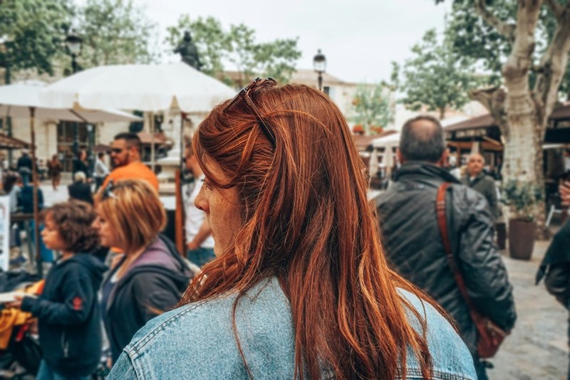 Red-haired woman in a denim jacket standing in a crowded outdoor market.