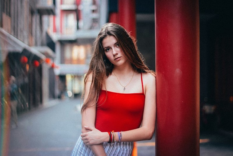 Young woman in a red top leaning beside a red column on a city street.