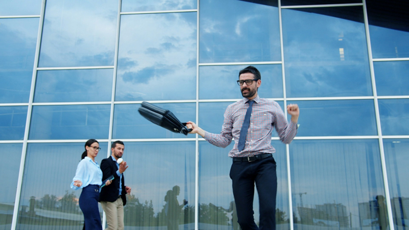 A businessman holding a suitcase jumps outside an office building, with two people watching in the background