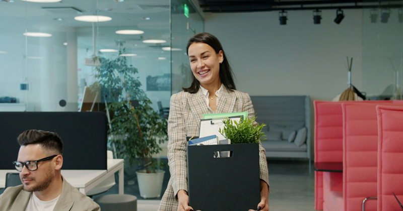 A female employee smiles while walking through an office, holding a box of her belongings