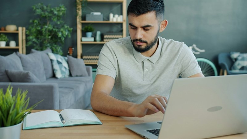A representation of a man working on a laptop and taking notes in a notebook at home.