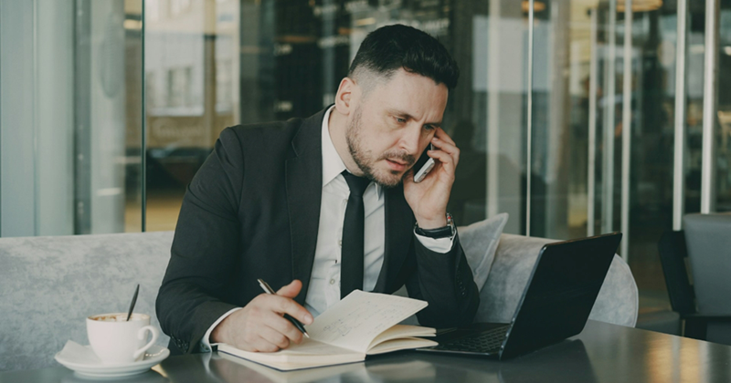 A male employee sits with his laptop while talking on his cell phone and writing in a notebook