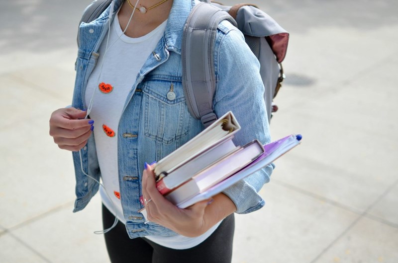 A high school student in a jean jacket and a white t-shirt carries books and listens to music while she walks the campus.