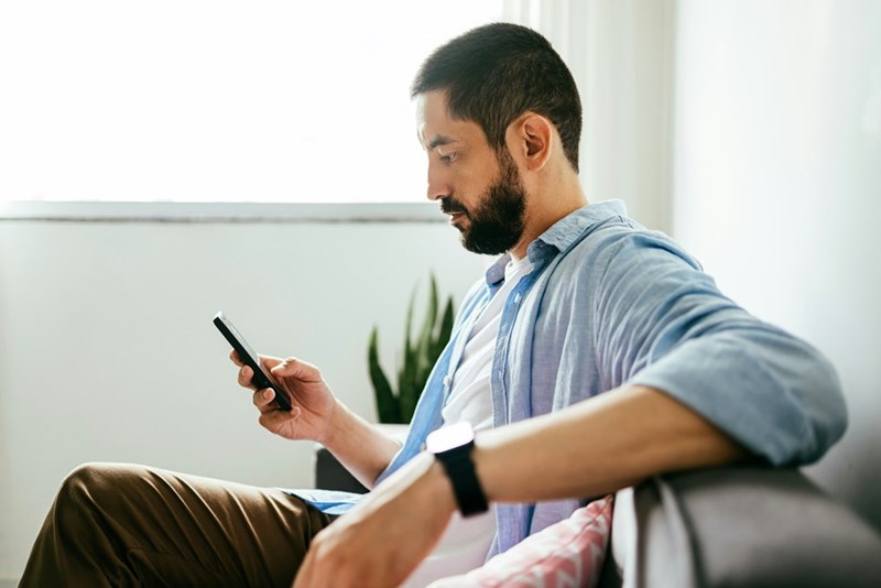 Man sitting on a couch and looking at his phone in a bright room with a plant behind him.