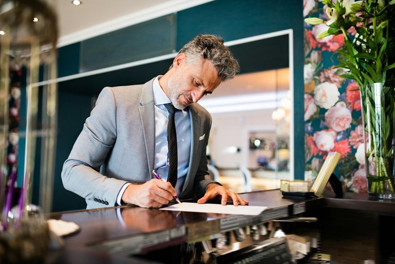 A man in a suit grey signs papers at the front desk of a very fancy hotel.