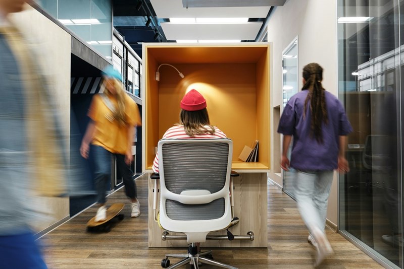 A worker in a red hat sits at a desk while others move around her