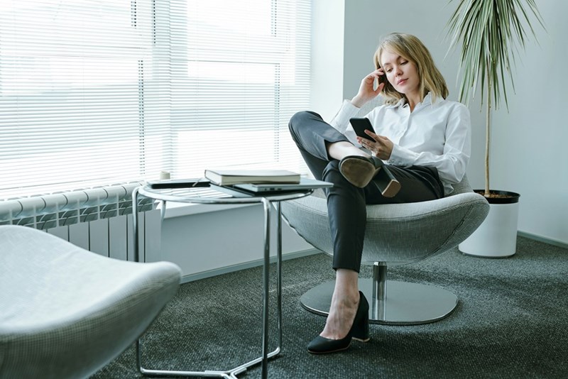 Young female employee sitting in an armchair by a large office window and scrolling on her phone, presented by a model.