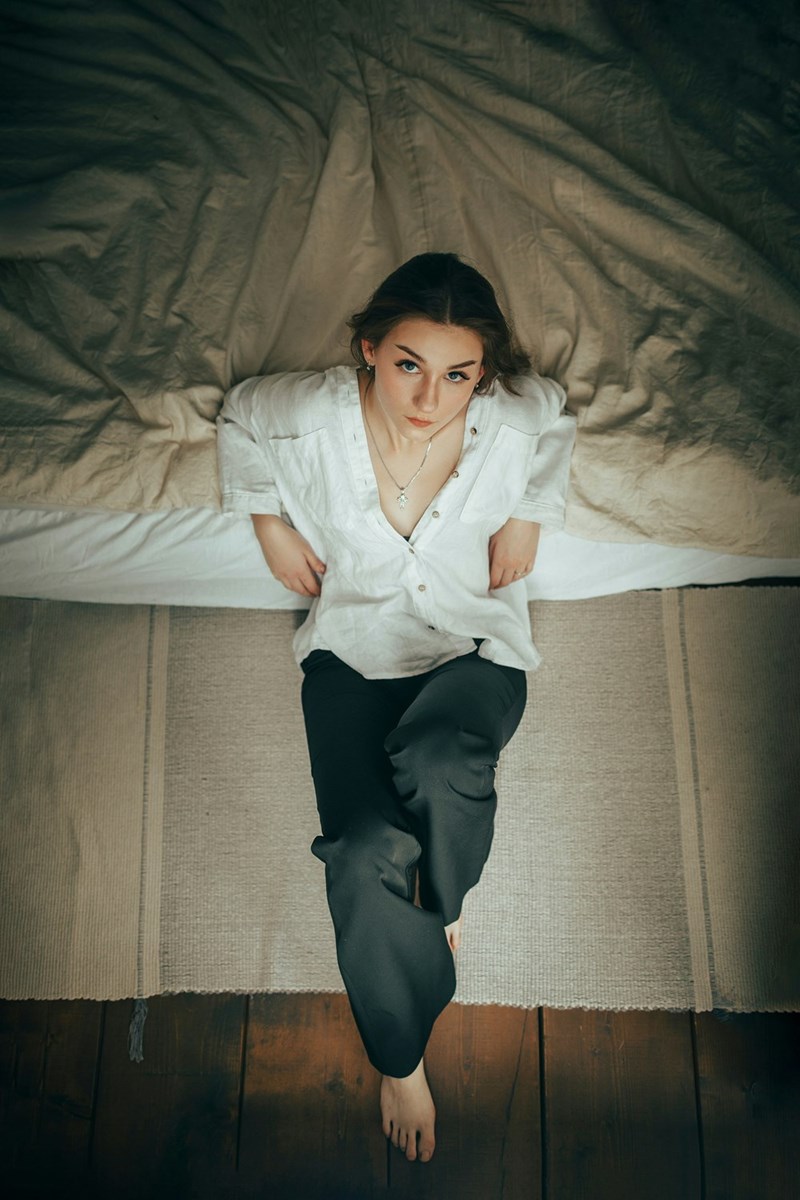 Woman sitting on the edge of a bed, looking up beside rumpled bedding and a wooden floor.