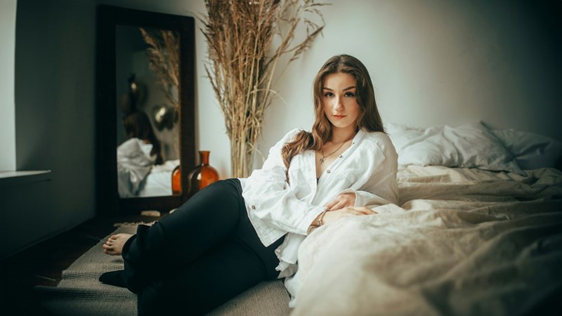Woman sitting beside a bed in a white shirt and black pants, with a mirror and rumpled bedding nearby.