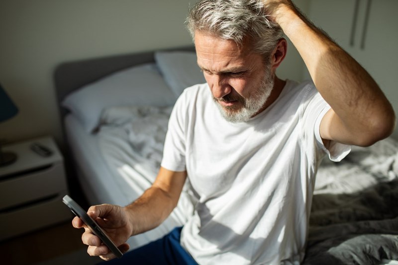 Man sitting on a bed holding a phone, with one hand in his hair in a sunlit bedroom.