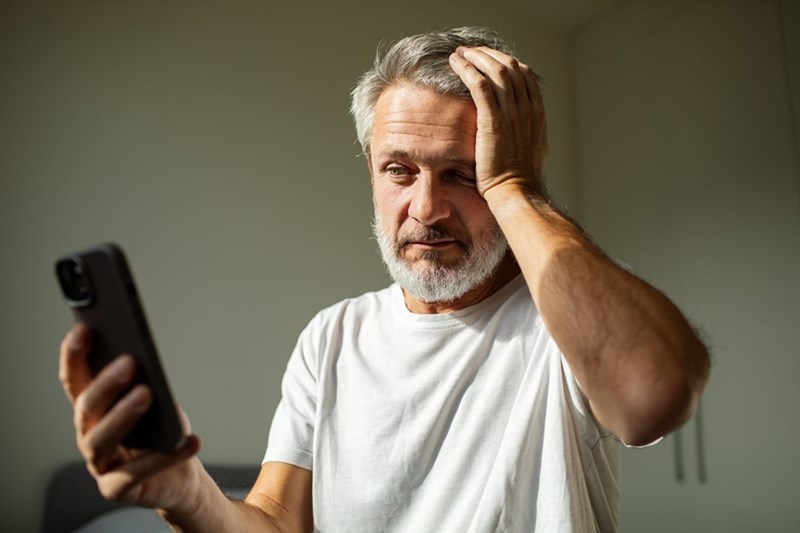 Man in a white T-shirt holding a phone with his hand on his head in a sunlit bedroom.