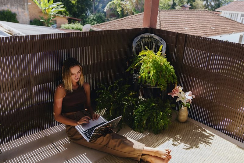 Woman sitting on a sunny balcony with a laptop, plants, privacy screen, and rooftops nearby.